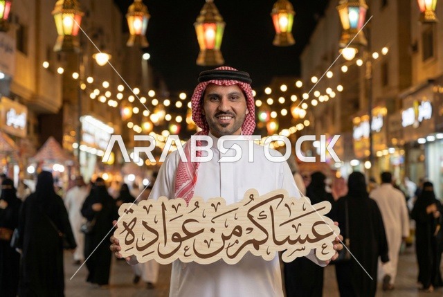 انوار الزينة الرمضانية المعلقة في الشوارع ليلا، مصابيح معدنية مضيئة، احبال الانارة، رجل عربي خليجي سعودي يرتدي الثوب التقليدي والشماغ ينظر الى الكاميرا بايماءات السعاده يحمل عبارة عساكم من عوادة ، فوانيس وزينة رمضان،  اجواء رمضانية