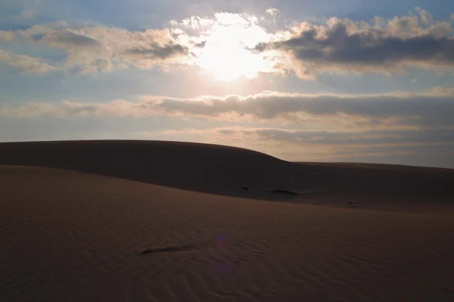 صحراء الدمر في مدينة صلالة وقت العصرية، سلطنة عُمان، مشهد صحراوي هادئ تلامسه ألوان الغروب، كثبان رملية تمتد تحت ضوء الشمس الذهبي، تباين ساحر بين الصحراء والسماء، أجواء طبيعية تعكس الطبيعة العُمانية في لحظات المساء.