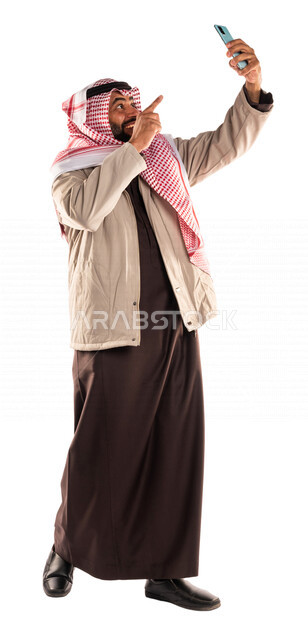 بورتريه لرجل عربي خليجي سعودي يرتدي ملابس الشتاء الدافئة ويشير الى الهاتف باصبع السبابة، التقاط صورة تذكارية من خلال الهاتف المحمول، صور تذكارية، صور سيلفي، كاميرا امامية، مكالمة فيديو ، بث فيديو مباشر، ايماءات وجه ويدين مبتسمة، صورة الجسم بالطول الكامل،