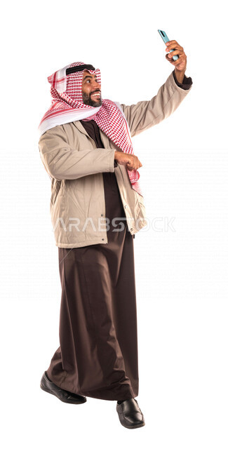 بورتريه لرجل عربي خليجي سعودي يرتدي ملابس الشتاء الدافئة، التقاط صورة تذكارية من خلال الهاتف المحمول، مكالمة فيديو ، بث فيديو مباشر، صور تذكارية، صور سيلفي، كاميرا امامية، ايماءات وجه ويدين مبتسمة، صورة الجسم بالطول الكامل، اجواء شتوية، خلفية بيضاء