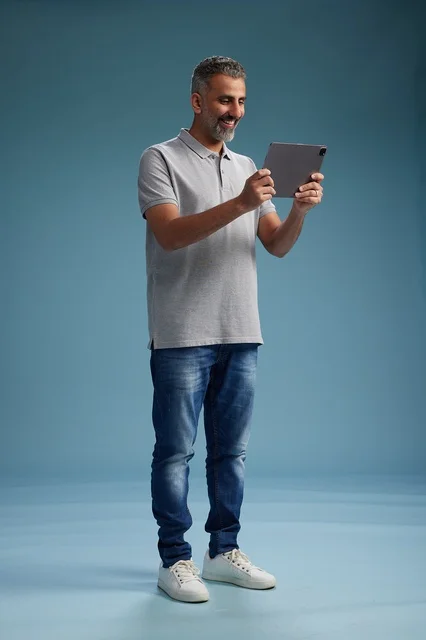 A Saudi Arabian Gulf man wearing casual attire is holding a tablet in his hand, with various gestures and movements, full-body, against a blue background.