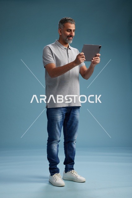 A Saudi Arabian Gulf man wearing casual attire is holding a tablet in his hand, with various gestures and movements, full-body, against a blue background.