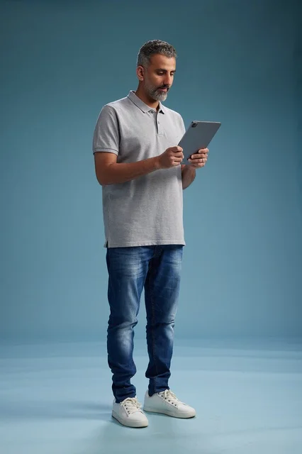 A Saudi Arabian Gulf man wearing casual attire is holding a tablet in his hand, with various gestures and movements, full-body, against a blue background.
