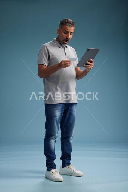 رجل عربي سعودي خليجي يرتدي الزي الكاجوال يقرأ بطاقة ويستخدم جهازًا لوحيًا, الدفع بالبطاقه الائتمانيه, مفهوم التسوق الاونلاين, خلفيه زرقاء, كامل الجسم