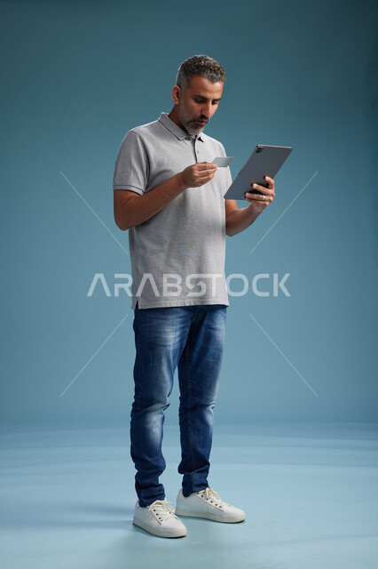 A Saudi Arabian Gulf Arab man in casual attire is reading a card and using a tablet, making a payment with a credit card, the concept of online shopping, blue background, full body.