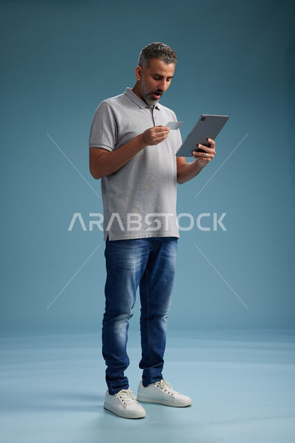 رجل عربي سعودي خليجي يرتدي الزي الكاجوال يقرأ بطاقة ويستخدم جهازًا لوحيًا, الدفع بالبطاقه الائتمانيه, مفهوم التسوق الاونلاين, خلفيه زرقاء, كامل الجسم