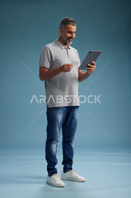 رجل عربي سعودي خليجي يرتدي الزي الكاجوال يقرأ بطاقة ويستخدم جهازًا لوحيًا, الدفع بالبطاقه الائتمانيه, مفهوم التسوق الاونلاين, خلفيه زرقاء, كامل الجسم