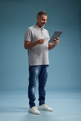 A Saudi Arabian Gulf Arab man in casual attire is reading a card and using a tablet, making a payment with a credit card, the concept of online shopping, blue background, full body.