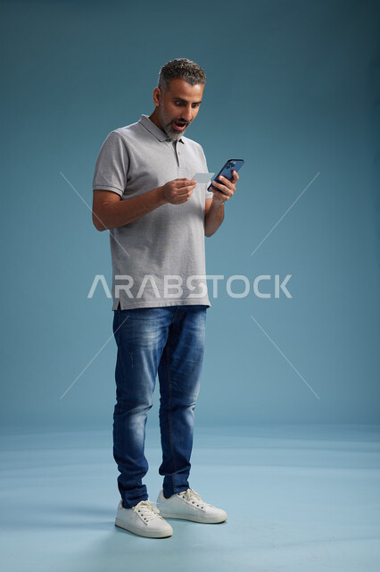 رجل عربي سعودي خليجي يرتدي الزي الكاجوال يقوم باجراء مكالمه هاتفيه ويحمل بطاقه بيضاء فارغه, كرت تعريفي , الجسم بالطول اكامل, خلفيه زرقاء