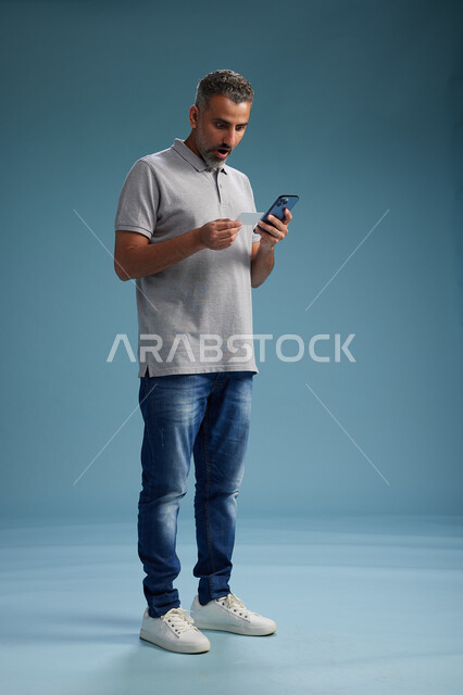 رجل عربي سعودي خليجي يرتدي الزي الكاجوال يقوم باجراء مكالمه هاتفيه ويحمل بطاقه بيضاء فارغه, كرت تعريفي , الجسم بالطول اكامل, خلفيه زرقاء