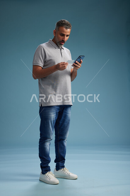 رجل عربي سعودي خليجي يرتدي الزي الكاجوال يقوم باجراء مكالمه هاتفيه ويحمل بطاقه بيضاء فارغه, كرت تعريفي , الجسم بالطول اكامل, خلفيه زرقاء