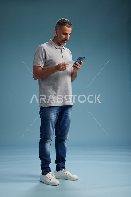 رجل عربي سعودي خليجي يرتدي الزي الكاجوال يقوم باجراء مكالمه هاتفيه ويحمل بطاقه بيضاء فارغه, كرت تعريفي , الجسم بالطول اكامل, خلفيه زرقاء