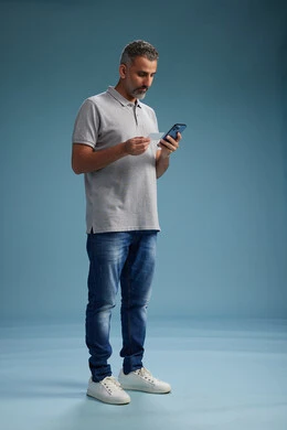 A Saudi Arabian Gulf Arab man dressed in casual attire is making a phone call while holding a blank white card, an identification card, with a full-length body, against a blue background.