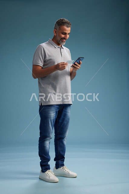رجل عربي سعودي خليجي يرتدي الزي الكاجوال يقوم باجراء مكالمه هاتفيه ويحمل بطاقه بيضاء فارغه, كرت تعريفي , الجسم بالطول اكامل, خلفيه زرقاء