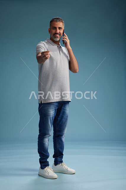 رجل عربي سعودي خليجي يرتدي الزي الكاجوال يقوم باجراء مكالمه هاتفيه ويحمل بطاقه بيضاء فارغه, كرت تعريفي , الجسم بالطول اكامل, خلفيه زرقاء