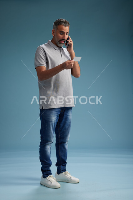 رجل عربي سعودي خليجي يرتدي الزي الكاجوال يقوم باجراء مكالمه هاتفيه ويحمل بطاقه بيضاء فارغه, كرت تعريفي , الجسم بالطول اكامل, خلفيه زرقاء