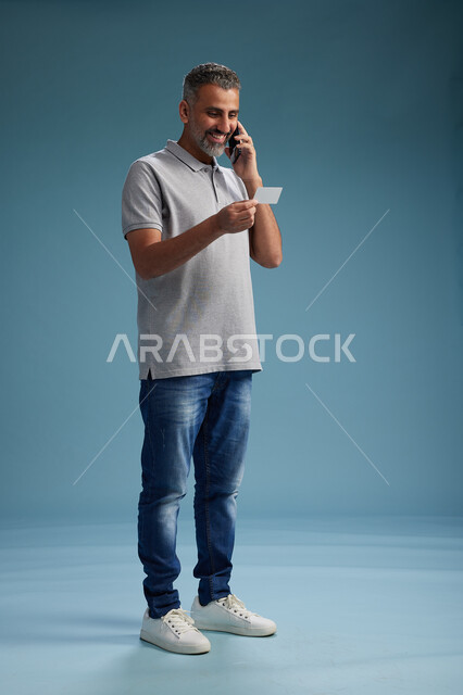 رجل عربي سعودي خليجي يرتدي الزي الكاجوال يقوم باجراء مكالمه هاتفيه ويحمل بطاقه بيضاء فارغه, كرت تعريفي , الجسم بالطول اكامل, خلفيه زرقاء