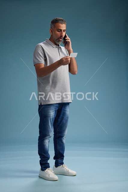 رجل عربي سعودي خليجي يرتدي الزي الكاجوال يقوم باجراء مكالمه هاتفيه ويحمل بطاقه بيضاء فارغه, كرت تعريفي , الجسم بالطول اكامل, خلفيه زرقاء