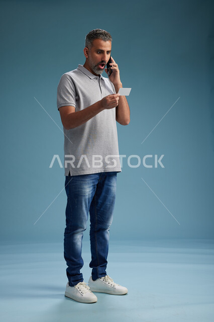 رجل عربي سعودي خليجي يرتدي الزي الكاجوال يقوم باجراء مكالمه هاتفيه ويحمل بطاقه بيضاء فارغه, كرت تعريفي , الجسم بالطول اكامل, خلفيه زرقاء
