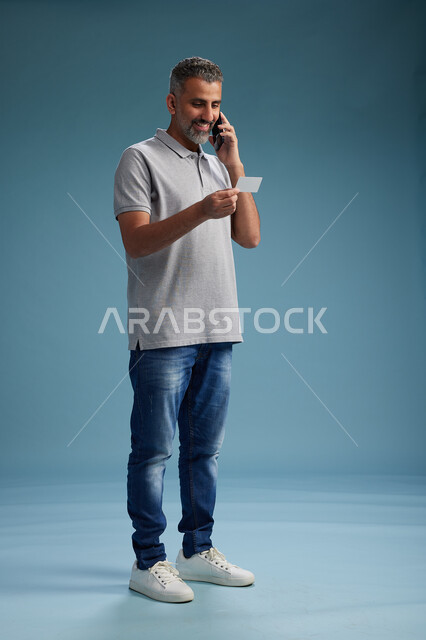 رجل عربي سعودي خليجي يرتدي الزي الكاجوال يقوم باجراء مكالمه هاتفيه ويحمل بطاقه بيضاء فارغه, كرت تعريفي , الجسم بالطول اكامل, خلفيه زرقاء