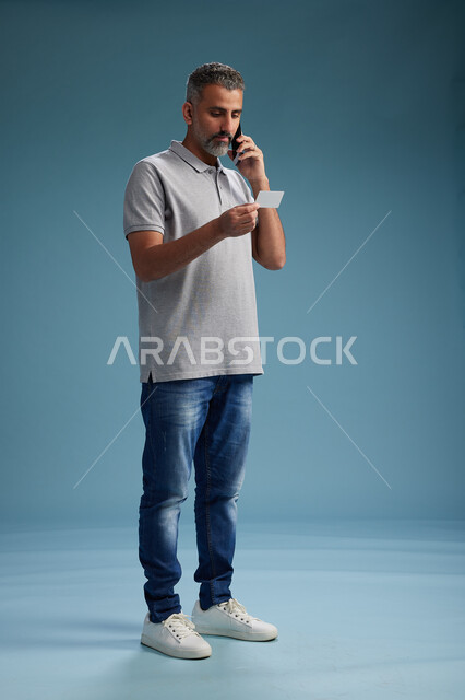 رجل عربي سعودي خليجي يرتدي الزي الكاجوال يقوم باجراء مكالمه هاتفيه ويحمل بطاقه بيضاء فارغه, كرت تعريفي , الجسم بالطول اكامل, خلفيه زرقاء
