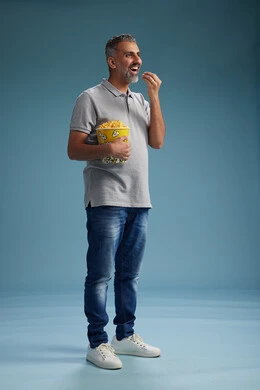 A Saudi Arabian Gulf Arab man wearing casual attire holding a bucket of popcorn, enjoying eating popcorn, full body with a blue background.