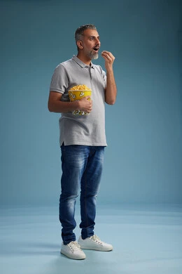 A Saudi Arabian Gulf Arab man wearing casual attire holding a bucket of popcorn, enjoying eating popcorn, full body with a blue background.