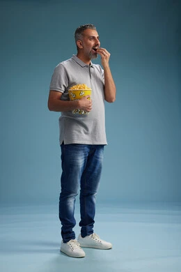 A Saudi Arabian Gulf Arab man wearing casual attire holding a bucket of popcorn, enjoying eating popcorn, full body with a blue background.