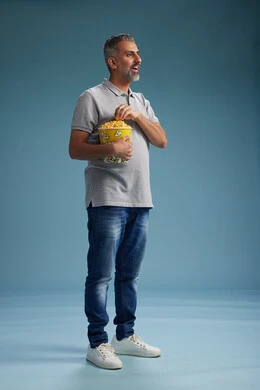 A Saudi Arabian Gulf Arab man wearing casual attire holding a bucket of popcorn, enjoying eating popcorn, full body with a blue background.