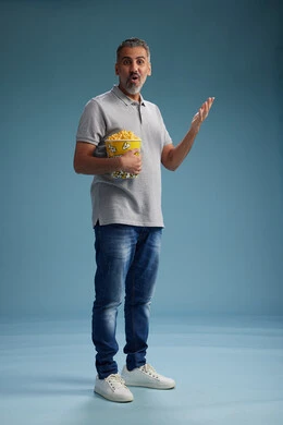 A Saudi Arabian Gulf Arab man wearing casual attire holding a bucket of popcorn, enjoying eating popcorn, full body with a blue background.