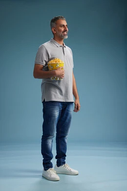 A Saudi Arabian Gulf Arab man wearing casual attire holding a bucket of popcorn, enjoying eating popcorn, full body with a blue background.