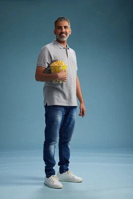 A Saudi Arabian Gulf Arab man wearing casual attire holding a bucket of popcorn, enjoying eating popcorn, full body with a blue background.