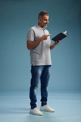 A Saudi Arabian Gulf Arab man wearing a gray shirt and jeans is reading a book about the concept of learning and culture, full body, with a blue background.