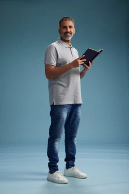 رجل عربي سعودي خليجي يرتدي قميص رمادي وبنطال جينز يقرأ كتابًا, مفهوم التعلم والثقافه, كامل الجسم, خلفيه زرقاء