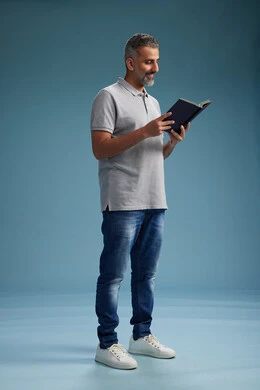 A Saudi Arabian Gulf Arab man wearing a gray shirt and jeans is reading a book about the concept of learning and culture, full body, with a blue background.