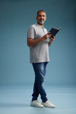 A Saudi Arabian Gulf Arab man wearing a gray shirt and jeans is reading a book about the concept of learning and culture, full body, with a blue background.