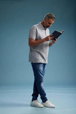 A Saudi Arabian Gulf Arab man wearing a gray shirt and jeans is reading a book about the concept of learning and culture, full body, with a blue background.