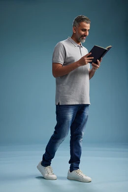 A Saudi Arabian Gulf Arab man wearing a gray shirt and jeans is reading a book about the concept of learning and culture, full body, with a blue background.