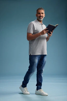 A Saudi Arabian Gulf Arab man wearing a gray shirt and jeans is reading a book about the concept of learning and culture, full body, with a blue background.