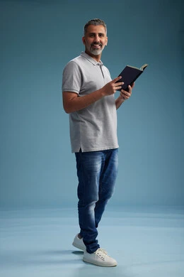 A Saudi Arabian Gulf Arab man wearing a gray shirt and jeans is reading a book about the concept of learning and culture, full body, with a blue background.