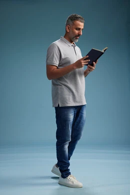 A Saudi Arabian Gulf Arab man wearing a gray shirt and jeans is reading a book about the concept of learning and culture, full body, with a blue background.