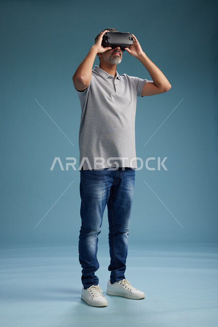 رجل عربي سعودي خليجي يرتدي الزي الكاجوال يستخدم نظارة الواقع الافتراضي, مشاهدة الافلام والفيديوز المسليه عبر الفي ار, كامل الجسم, خلفيه زرقاء