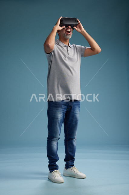 رجل عربي سعودي خليجي يرتدي الزي الكاجوال يستخدم نظارة الواقع الافتراضي, مشاهدة الافلام والفيديوز المسليه عبر الفي ار, كامل الجسم, خلفيه زرقاء