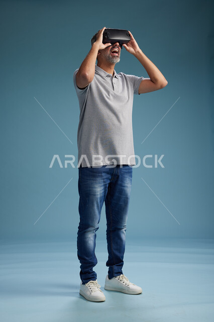 رجل عربي سعودي خليجي يرتدي الزي الكاجوال يستخدم نظارة الواقع الافتراضي, مشاهدة الافلام والفيديوز المسليه عبر الفي ار, كامل الجسم, خلفيه زرقاء