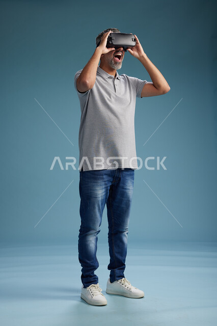 رجل عربي سعودي خليجي يرتدي الزي الكاجوال يستخدم نظارة الواقع الافتراضي, مشاهدة الافلام والفيديوز المسليه عبر الفي ار, كامل الجسم, خلفيه زرقاء