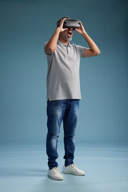 A Saudi Arabian Gulf Arab man wearing casual attire is using virtual reality glasses, watching movies and entertaining videos through VR, full body, with a blue background.