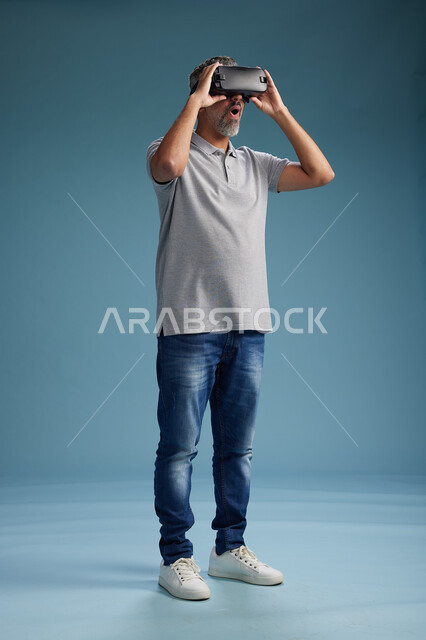 A Saudi Arabian Gulf Arab man wearing casual attire is using virtual reality glasses, watching movies and entertaining videos through VR, full body, with a blue background.