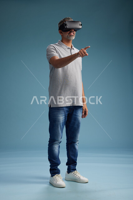 رجل عربي سعودي خليجي يرتدي الزي الكاجوال يستخدم نظارة الواقع الافتراضي, مشاهدة الافلام والفيديوز المسليه عبر الفي ار, كامل الجسم, خلفيه زرقاء