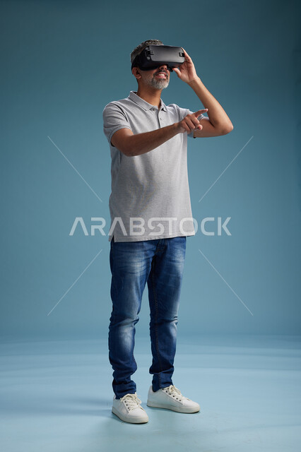 رجل عربي سعودي خليجي يرتدي الزي الكاجوال يستخدم نظارة الواقع الافتراضي, مشاهدة الافلام والفيديوز المسليه عبر الفي ار, كامل الجسم, خلفيه زرقاء