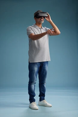 A Saudi Arabian Gulf Arab man wearing casual attire is using virtual reality glasses, watching movies and entertaining videos through VR, full body, with a blue background.